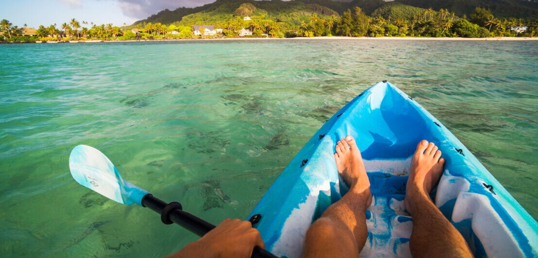 Nackte Beine on einem blauen Kanu auf dem Wasser vor tropischer Küstenlandschaft.