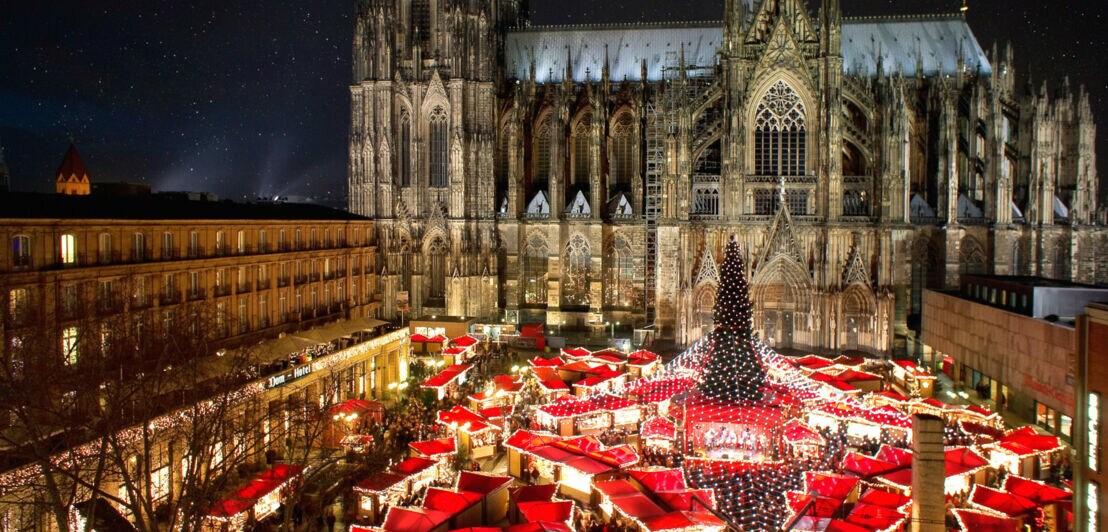 Beleuchteter Weihnachtsmarkt mit Tannenbaum am Kölner Dom bei Dunkelheit.