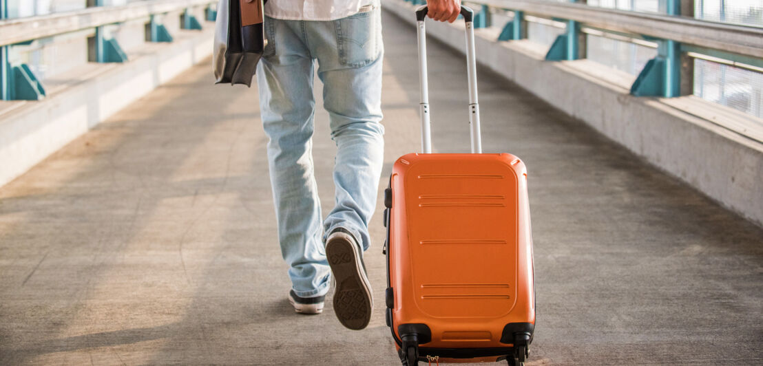 Rückansicht einer Person in Jeans, die einen orangefarbenen Hartschalenkoffer in einem Durchgang hinter sich herzieht.