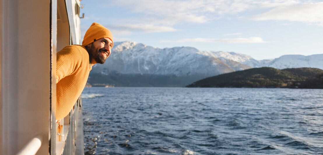 Ein lächelnder Mann mit gelbem Pullover und Mütze schaut von einem Schiff aufs Wasser vor schneebedeckter Berglandschaft.