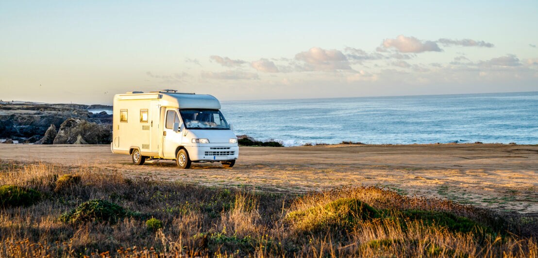 Ein Camper auf einer freien Fläche mit Sand an einer Küste bei Sonnenuntergang.