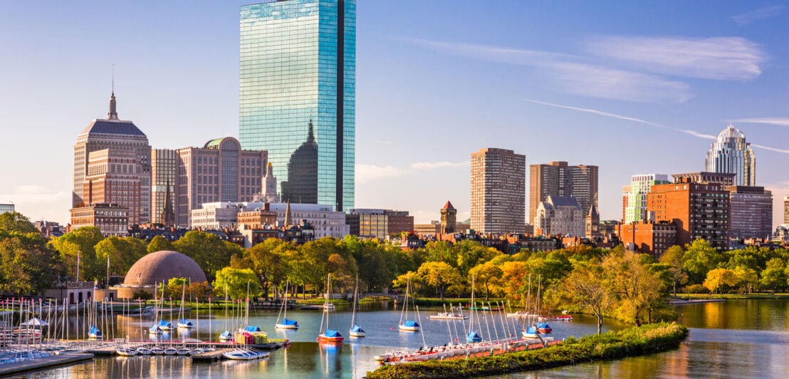 Skyline von Boston mit Parkanlage am Wasser mit Booten vor Hochhäusern im Hintergrund.