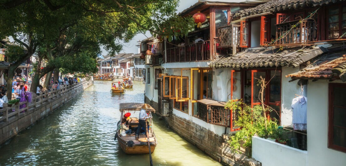 Boote mit Personen mit asiatischem Kegelhut auf einem Kanal entlang einer Häuserfront, links Menschenmenge auf einer Promenade.