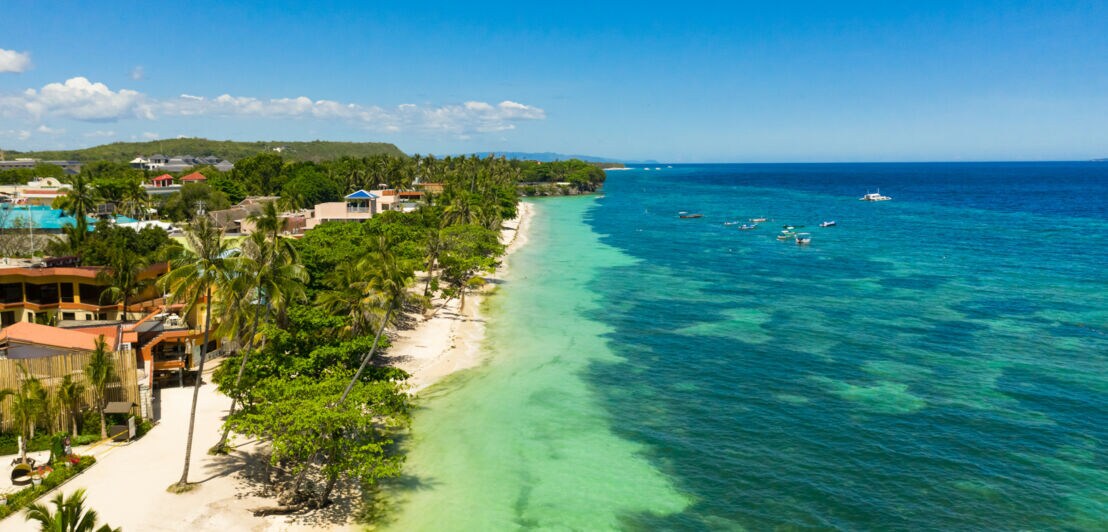 Bungalows auf einem palmengesäumten Sandstrand an türkisblauer Meeresküste.