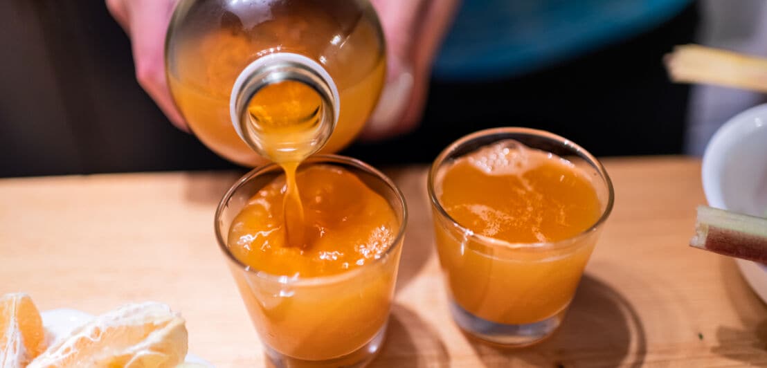 Ein orangefarbenes Getränk wird von einer Glasflasche in ein Glas gefüllt, daneben ein weiteres gefülltes Glas auf einem Holztisch.