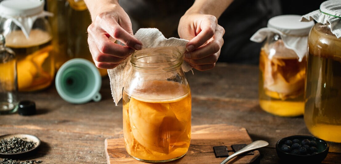 Zwei Hände bedecken mit einem Stofftuch ein Einmachglas mit Kombucha-Getränk samt Teepilz auf einem Holztisch, daneben weitere befüllte Einmachgläser.