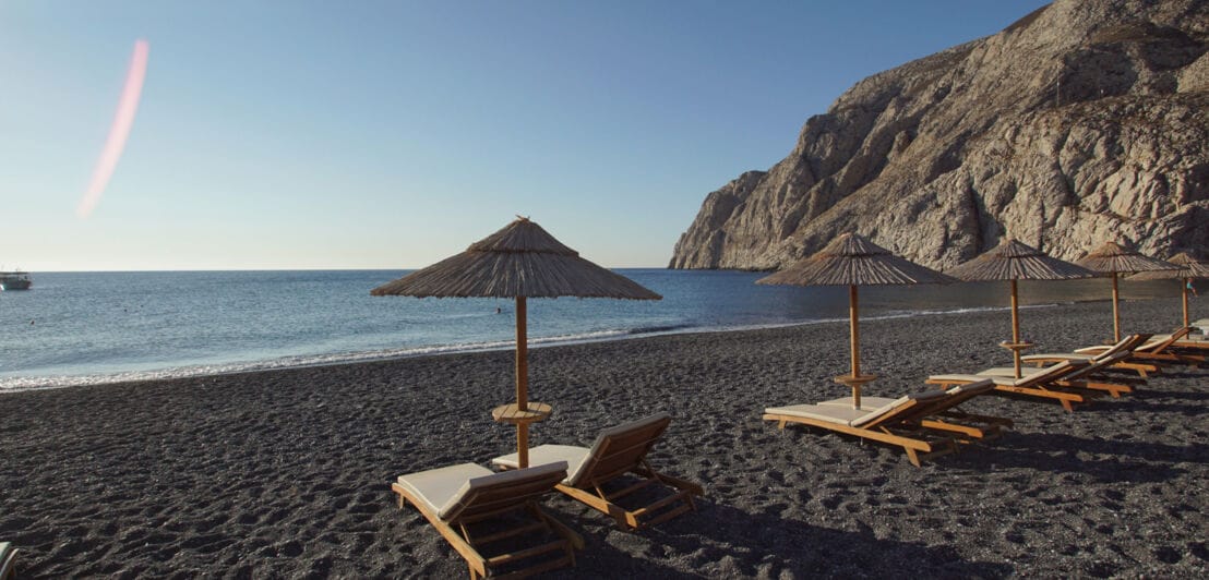 Liegen und Sonnenschirme an einem menschenleeren Strand mit schwarzem Sand in einer Bucht.