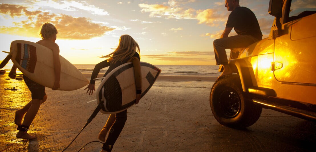 Rückansicht von zwei Kindern mit Surfbrettern neben einem sitzenden Mann auf der Motorhaube eines Jeeps am Strand bei Sonnenuntergang.