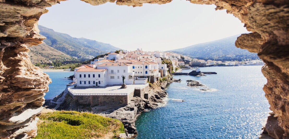 Blick auf eine Landzunge mit weißen Häusern an einer mediterranen Küste durch ein Loch in einem Felsblock.