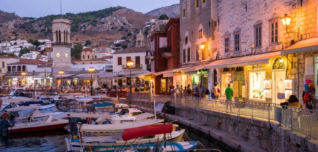 Fischerboote im Wasser entlang einer belebten Hafenpromenade in einer mediterranen Ortschaft am Abend.