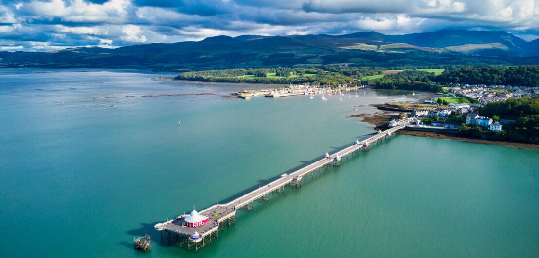 Langer Pier an einem Küstenort, im Hintergrund grüne Hügellandschaft.