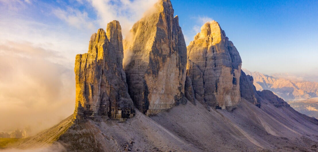 Blick auf drei markante Felstürme der Dolomiten im Sonnenschein, im Hintergrund blauer Himmel.