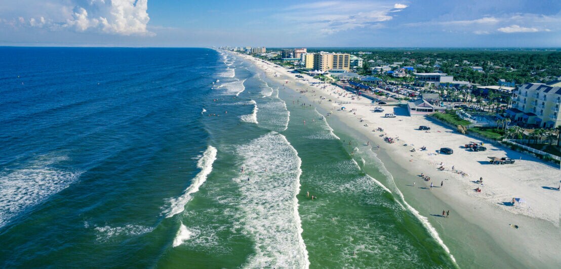 Stadt mit breitem Sandstrand am Meer mit Wellengang aus der Luftperspektive.