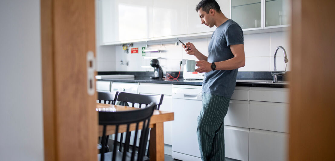 Ein Mann steht in einer Küche und hält in einer Hand eine Tasse und in der anderen ein Smartphone.