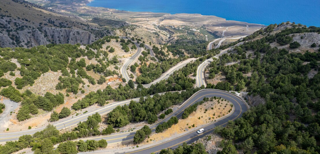 Ein Auto auf einer Serpentinenstraße in einer mediterranen Berglandschaft an einer Meeresküste.