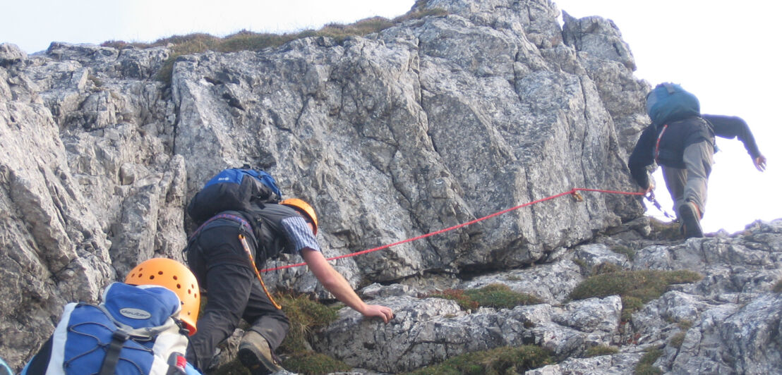 Drei Personen mit Kletterausrüstung, die einen steilen Felsen hochklettern, an dem ein Seil gespannt ist.