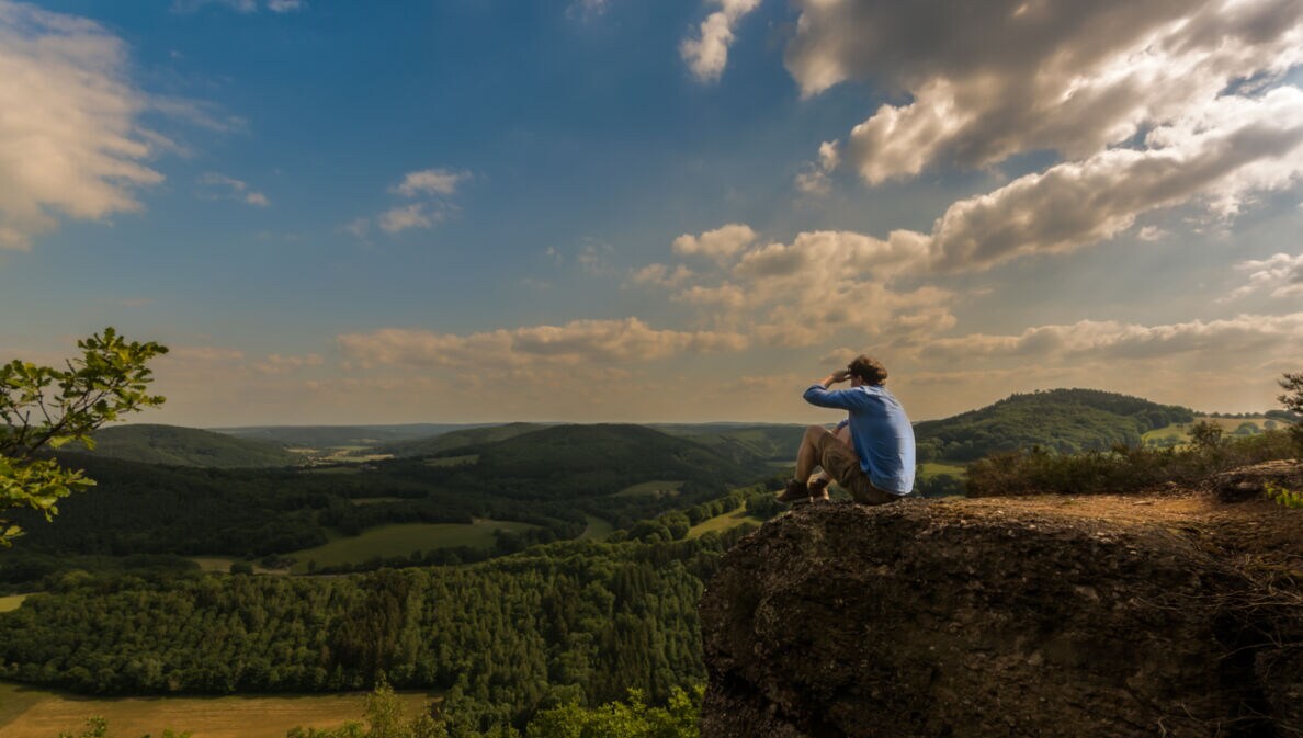 Ein Mann sitzt auf einem Felsen und blickt über die grüne Landschaft der Eifel