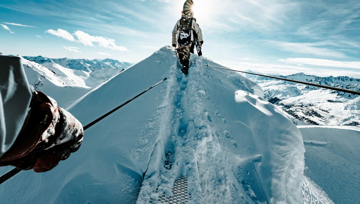 Ein:e Wintersportler:in mit Snowboard auf dem Rücken geht über einen schneebedeckten Berggipfel.