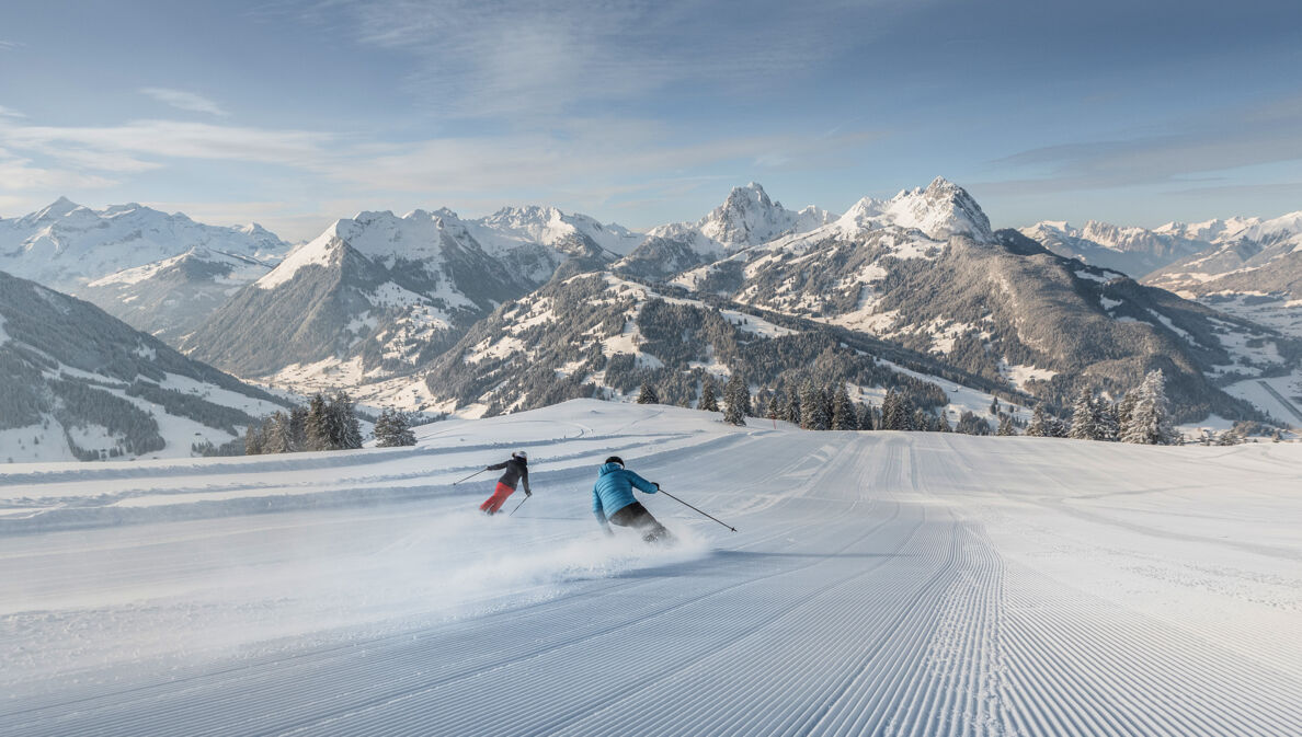 Rückansicht zweier Skifahrenden auf einer Piste vor Gipfelpanorama.