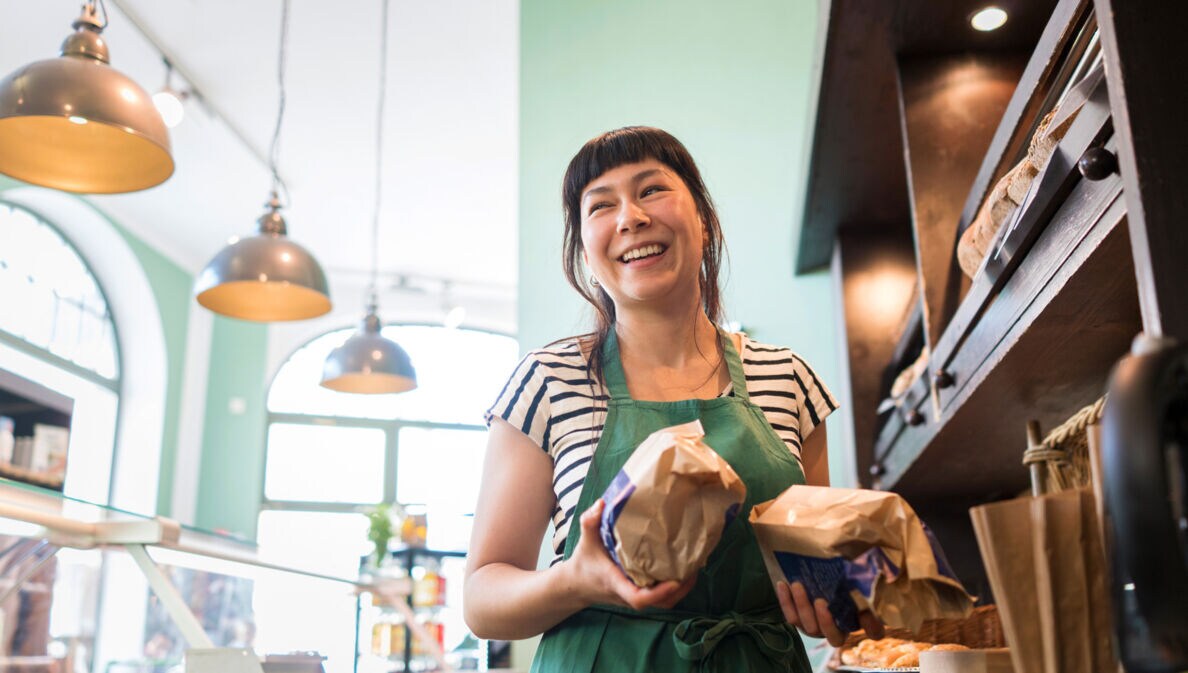 Eine Verkäuferin mit Backwaren in den Händen steht lachend hinter der Theke einer Bäckerei.