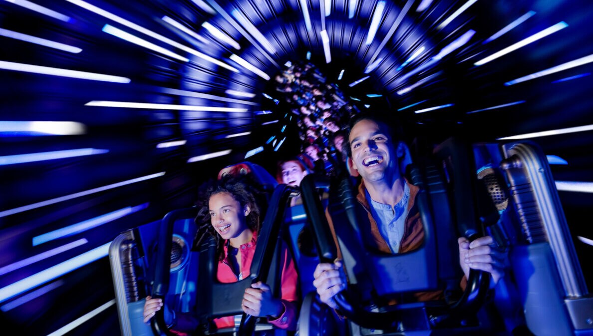Ein Mann und ein Mädchen fahren fröhlich im Wagen einer Achterbahn durch einen dunklen Tunnel mit Lichteffekten