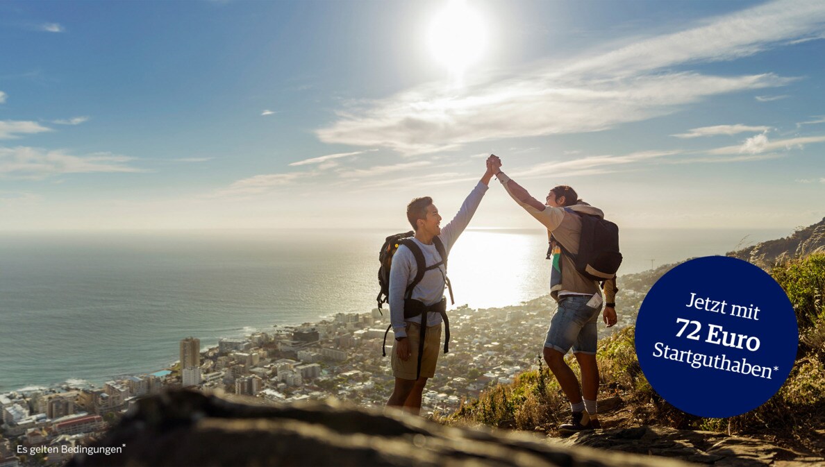 Zwei Wanderer mit Rucksäcken geben sich ein High Five auf einer Bergspitze, im Hintergrund eine Großstadt am Meer.