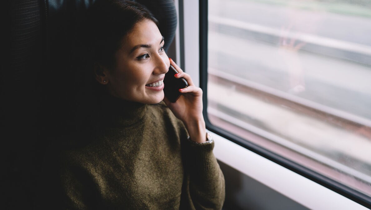 Lächelnde Frau telefoniert in fahrendem Zug und schaut aus dem Fenster