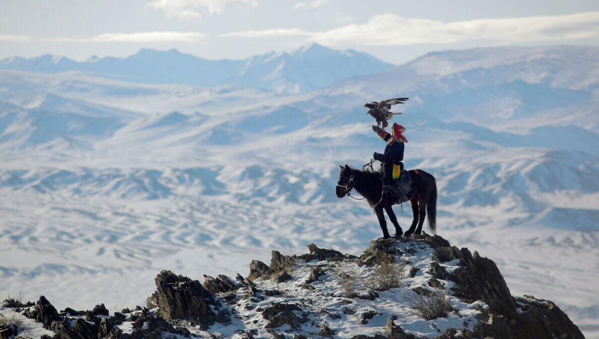 Ein Mann sitzt auf einem Pferd im Gebirge, auf seinem ausgestreckten Arm steht ein Adler.