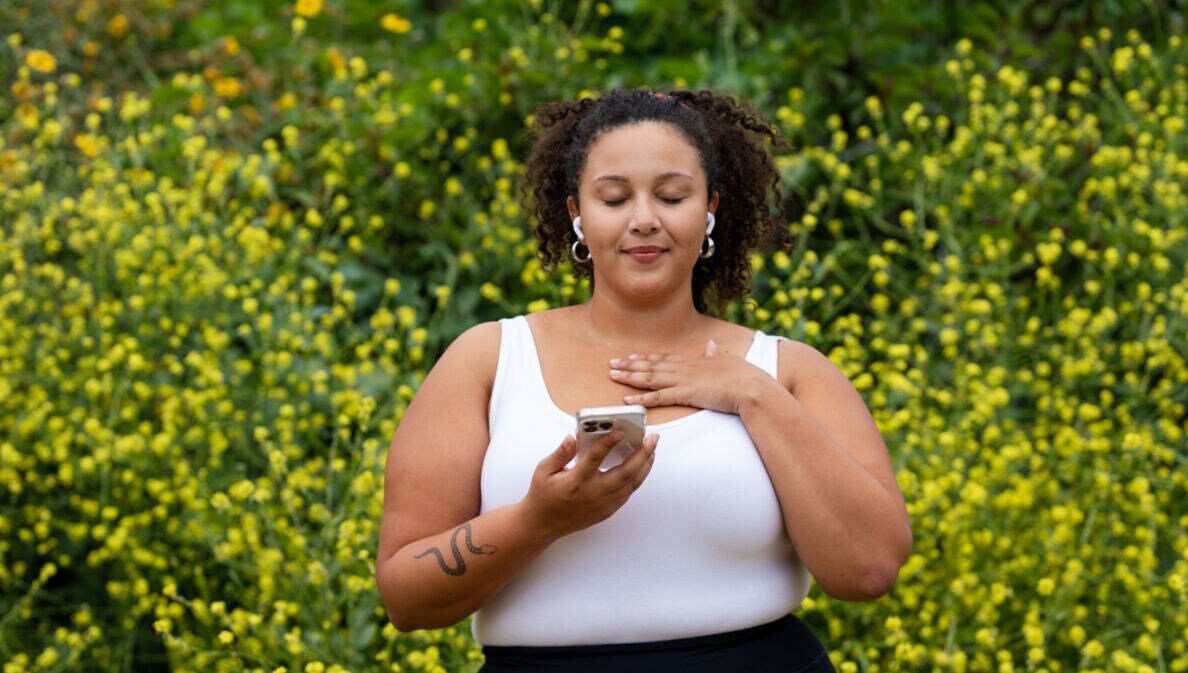 Eine Frau mit Smartphone in der Hand steht mit geschlossenen Augen in der Natur und meditiert.