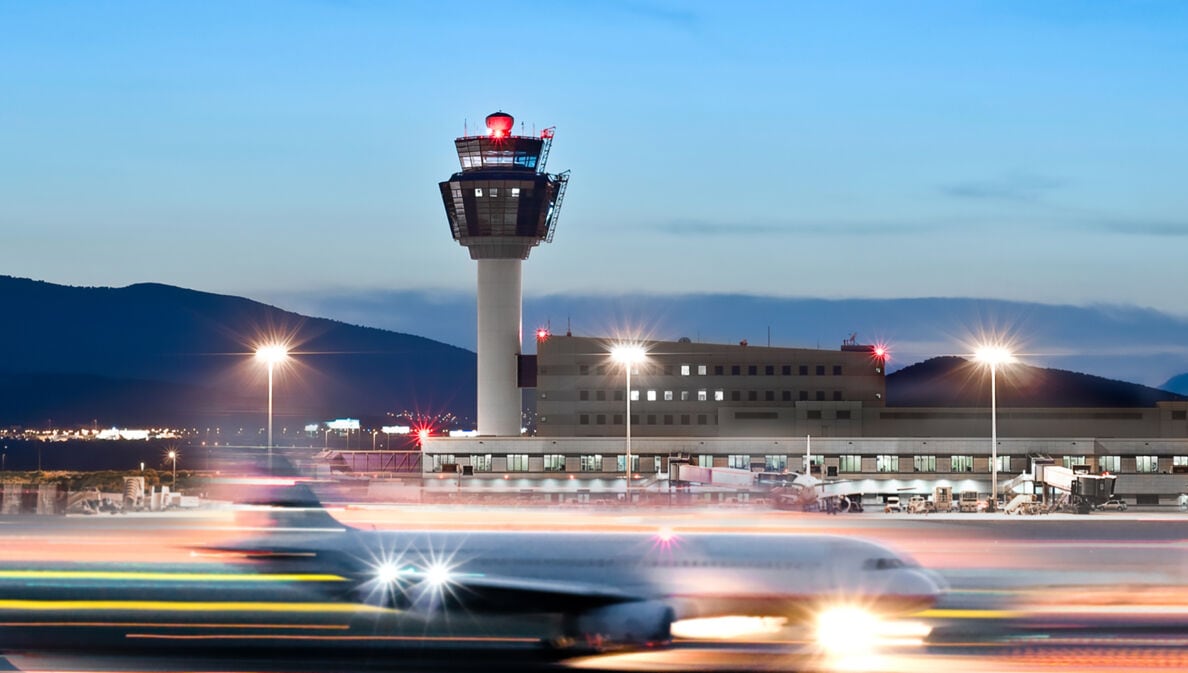 Ein Flugzeug in Bewegungsunschärfe auf dem Rollfeld vor einem Flughafenterminal mit Tower.
