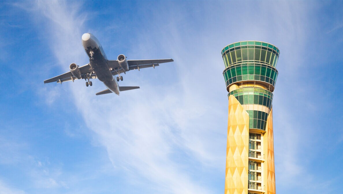 Untersicht eines Flugzeuges am blauen Himmel neben einem grün-gelben Tower. 