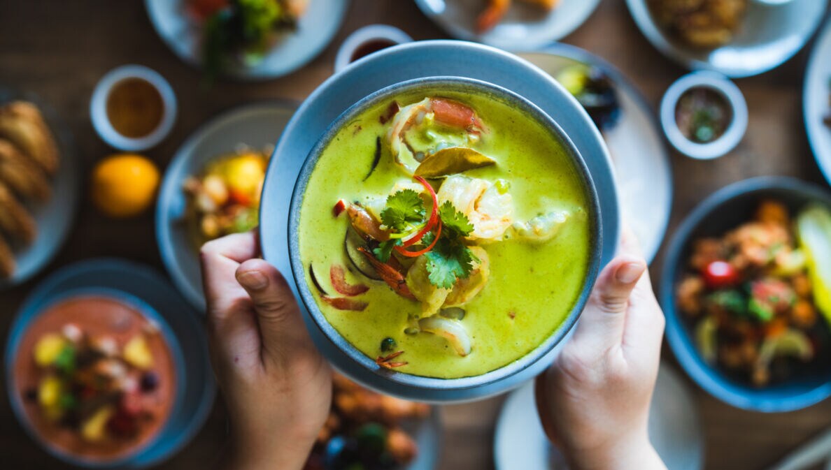 Zwei Hände halten eine Schale mit einem grünen Curry-Gericht über einen gedeckten Tisch mit vielen weiteren Speisen.