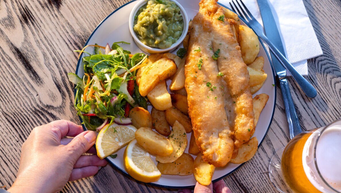 Eine Hand greift nach einem Teller Fish and Chips mit Beilagen auf einem Holztisch bei Sonnenschein.