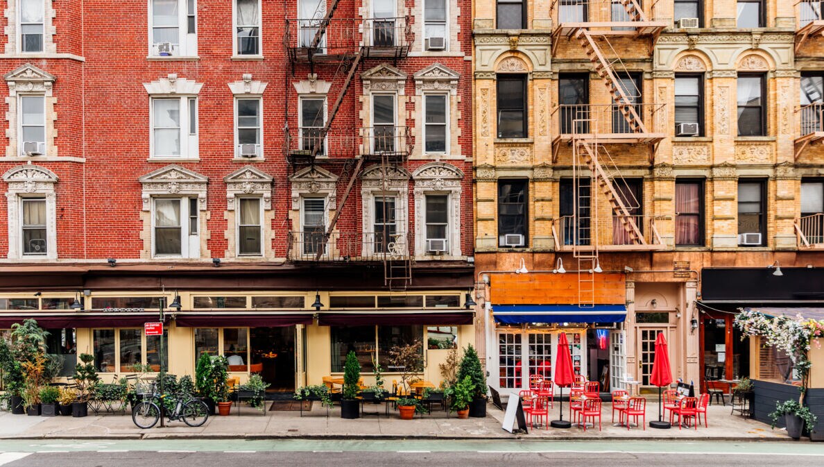 Geschlossene New Yorker Häuserfront mit Feuerleitern und Restaurants mit Außengastronomie.
