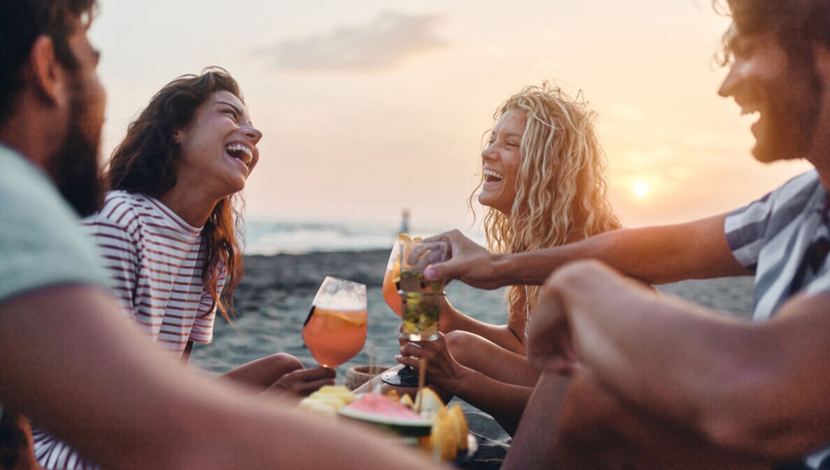 Eine Gruppe von vier fröhlichen Personen sitzt am Meer bei Sonnenuntergang mit Drinks und Snacks.