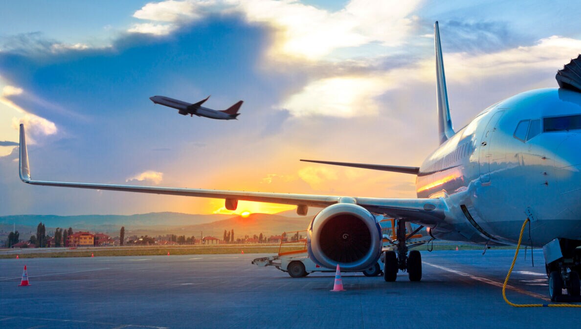 Ein Flugzeug am Himmel über einem geparkten Flugzeug am Flughafen vor einer hügeligen Landschaft bei Sonnenuntergang.