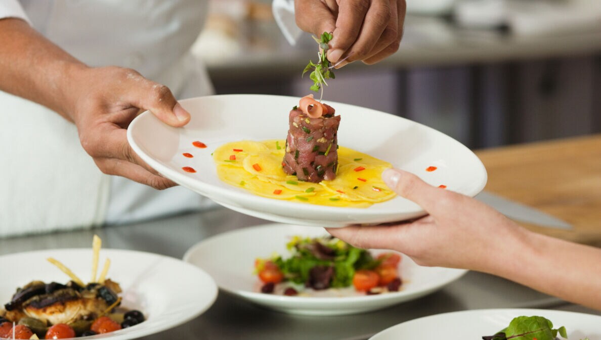Ein Koch garniert ein Gericht mit Thunfischtartar auf einem weißen Teller in einer Restaurantküche.