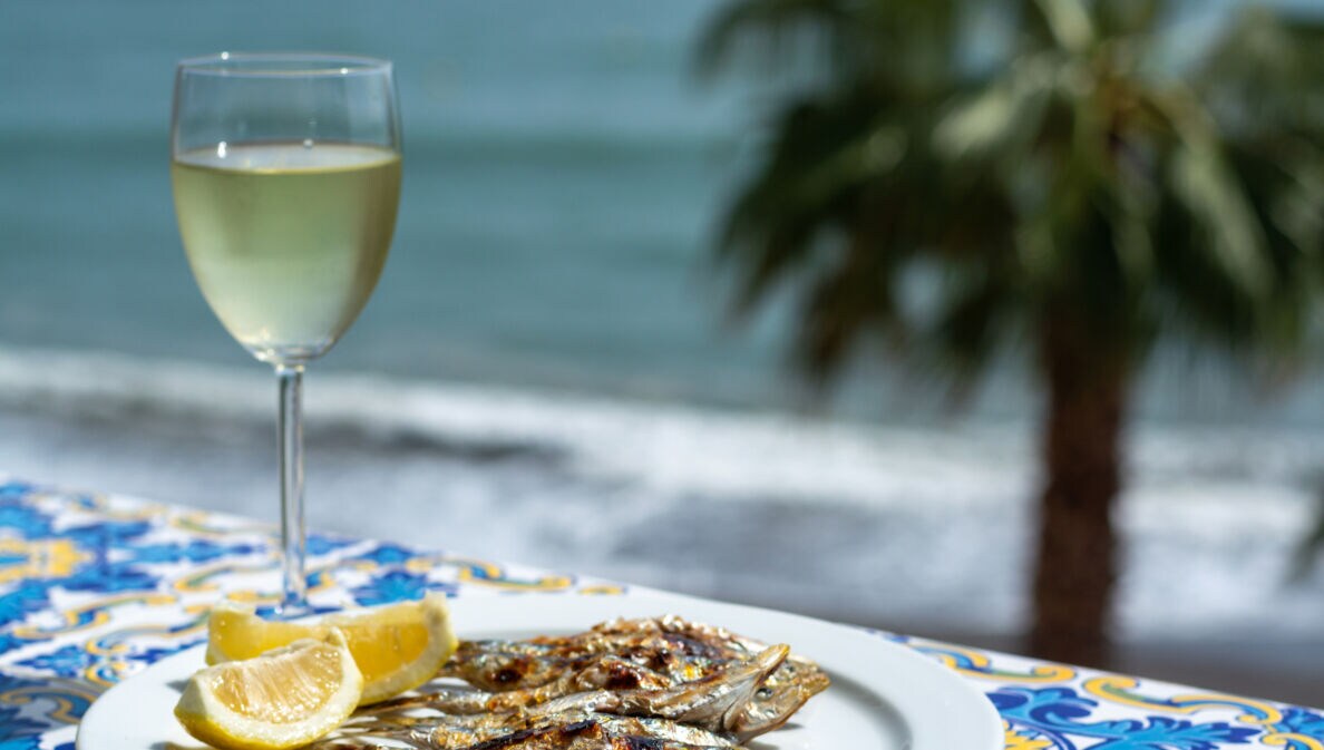 Gegrillte Sardinen auf einem weißen Teller neben einem Glas Weißwein, dahinter Meer mit Palme in Unschärfe.