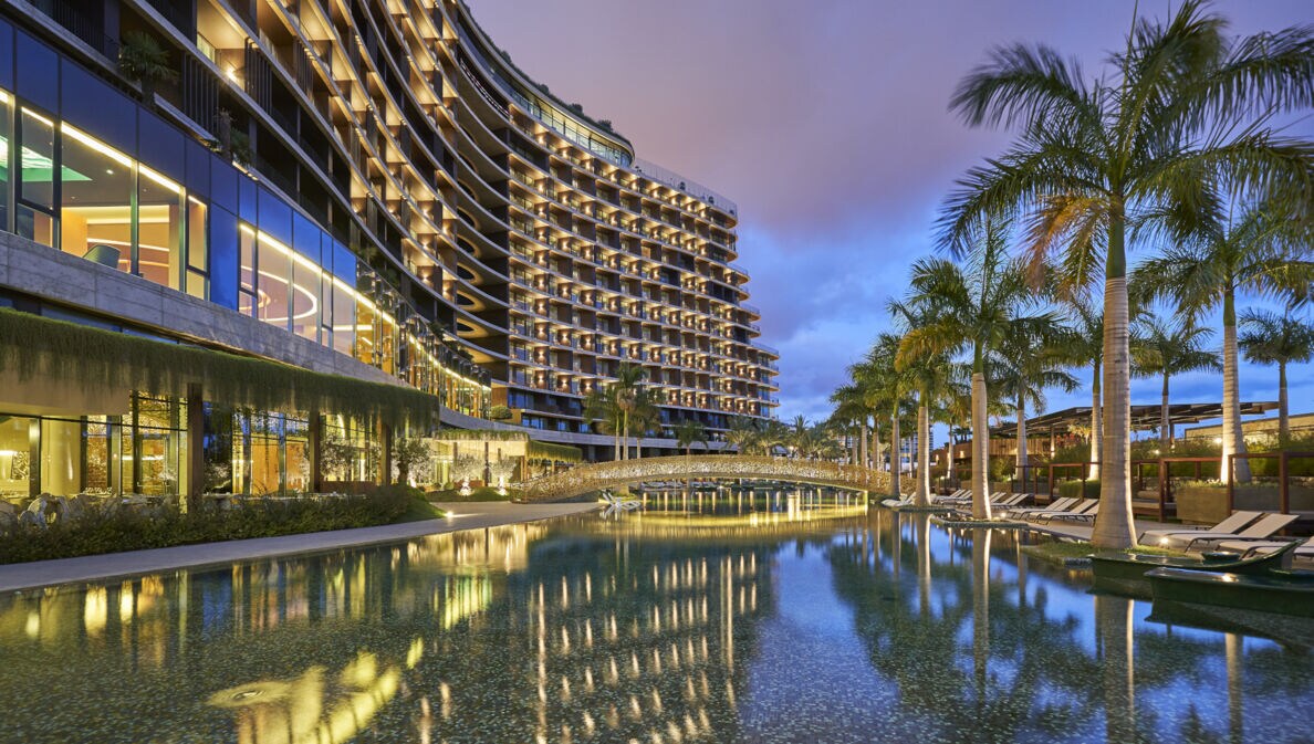 Blick auf ein beleuchtetes Luxushotel mit Palmen und Wasserbecken.