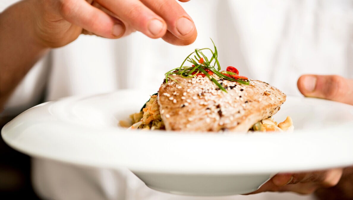 Eine Hand verfeinert ein Fleischgericht auf einem weißen Teller in der anderen Hand.