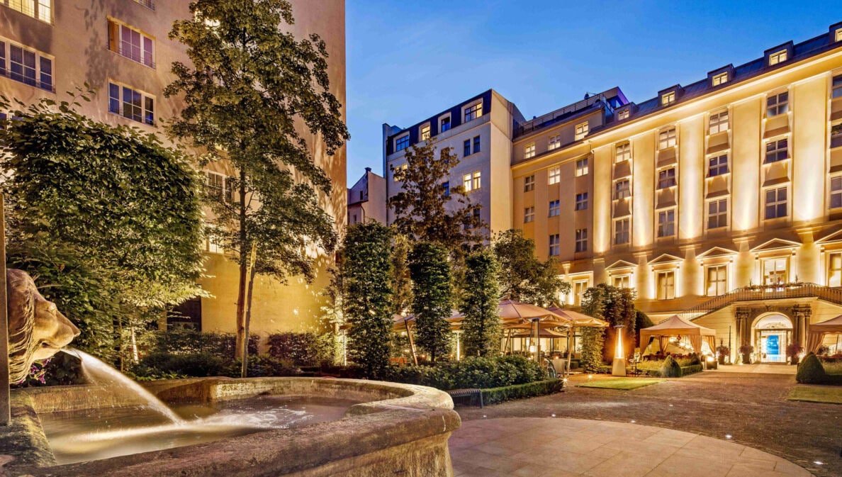 Imposantes, beleuchtetes Hotelgebäude mit barocker Fassade, im Vordergrund ein Springbrunnen mit Löwenkopf.