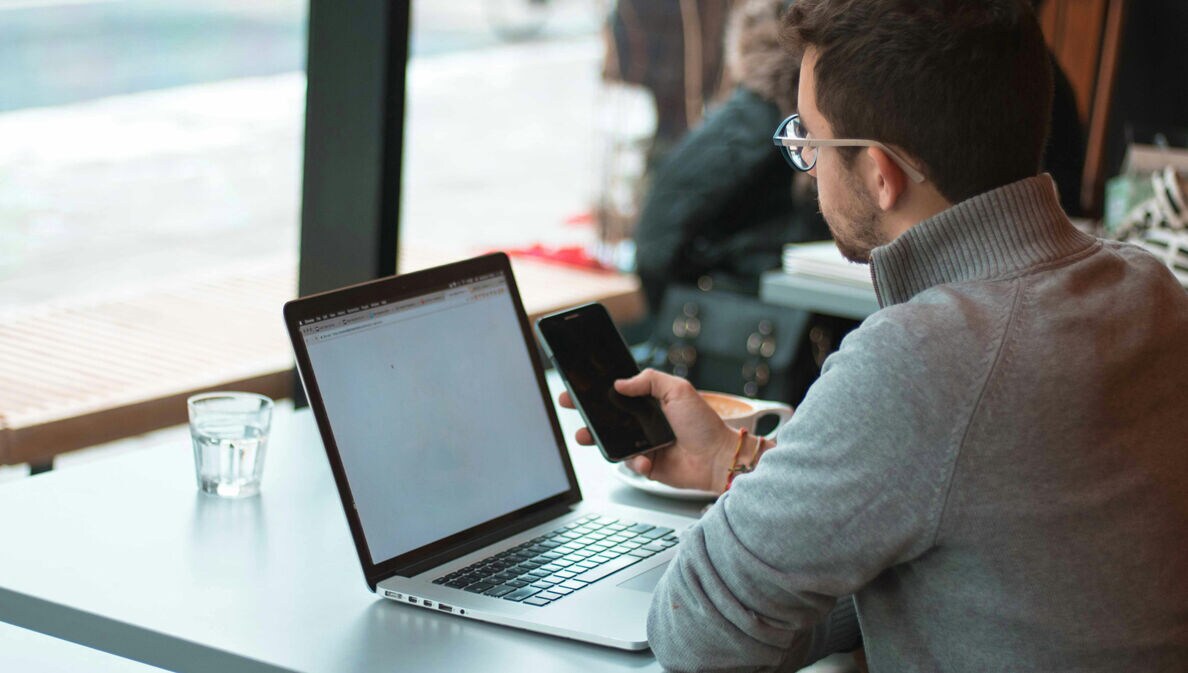 Mann sitzt mit Laptop und im Cafe und schat auf sein Smartphone