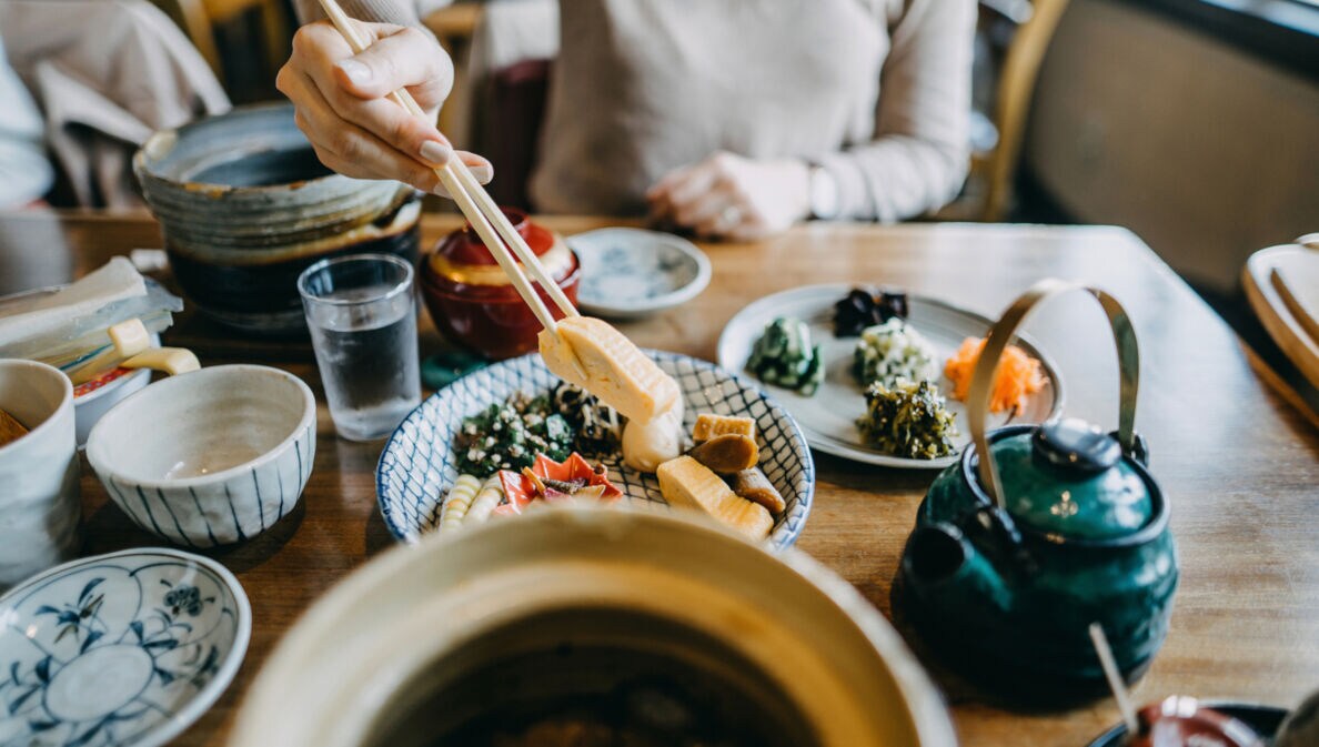 Auf einem Tisch stehen mehrere Schälchen mit verschiedenen asiatischen Gerichten. Eine unkenntliche Person hält mit Stäbchen etwas zu essen hoch.