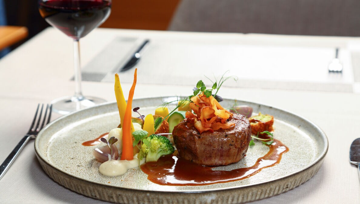 Ein Stück Steak an Gemüse, angerichtet auf einem Teller neben einem Glas Rotwein auf einem weiß gedeckten Tisch.