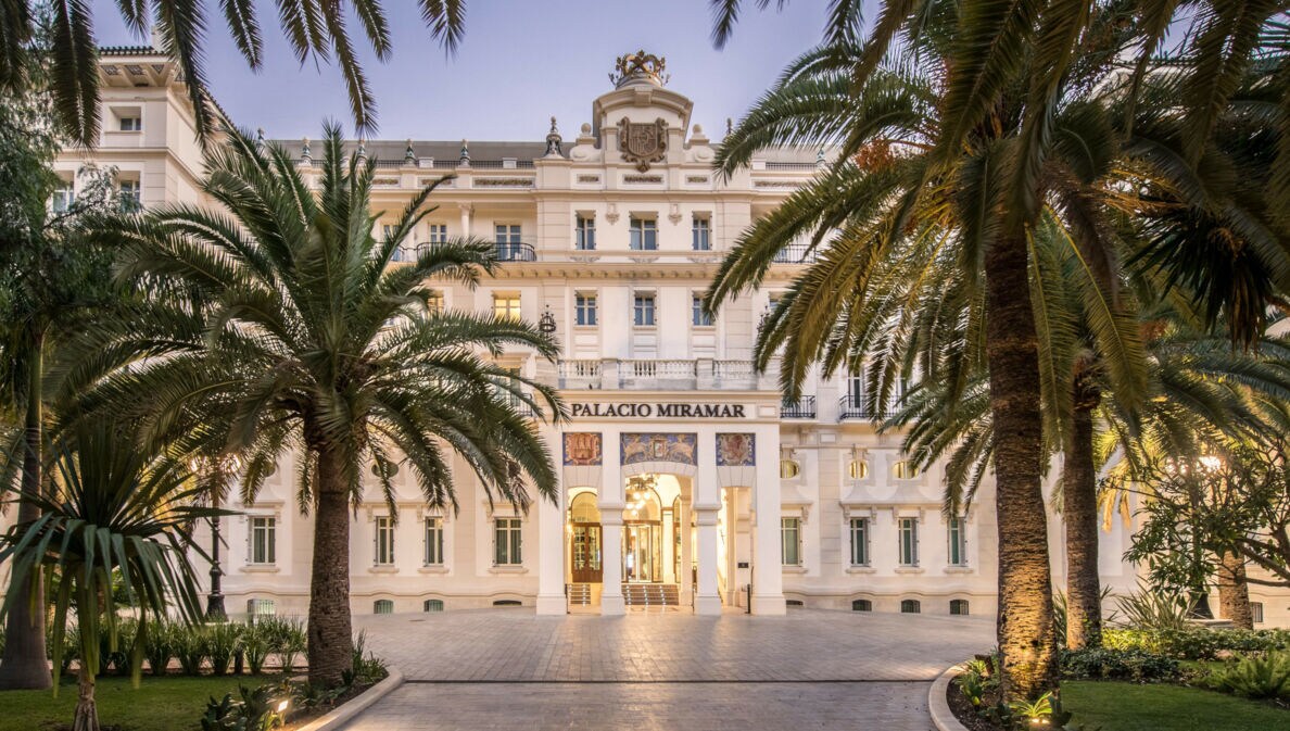 Imposante, helle Frontfassade eines Grandhotels mit Vorplatz, im Vordergrund Begrünung mit Palmen.