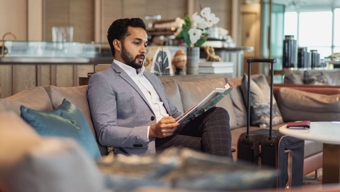 Ein elegant gekleideter Mann liest eine Zeitschrift auf einem Sofa in einer Flughafenlounge.