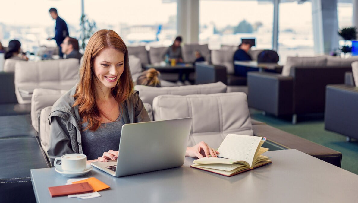 Eine lächelnde Frau arbeitet an ihrem Laptop an einem Tisch in einer Flughafen-Lounge.