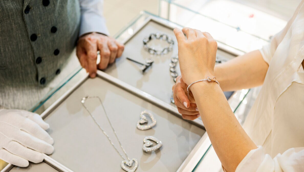 Der Arm einer Frau, die gerade ein Diamantarmband anzieht, da drunter ein Tisch mit mehreren Schmuckstücken.