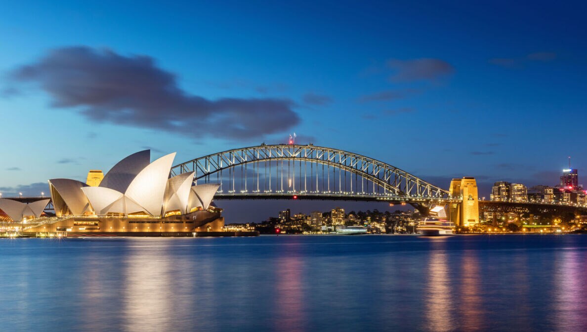 Sydney Harbour Bogenbrücke hinter dem Sydney Opernhaus am Wasser vor der erleuchteten Skyline Sydneys bei Nacht.