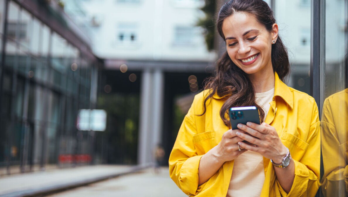 Eine Frau im gelben Hemd lehnt an einer Außenfassade und schaut lächelnd auf das Smartphone in ihren Händen.